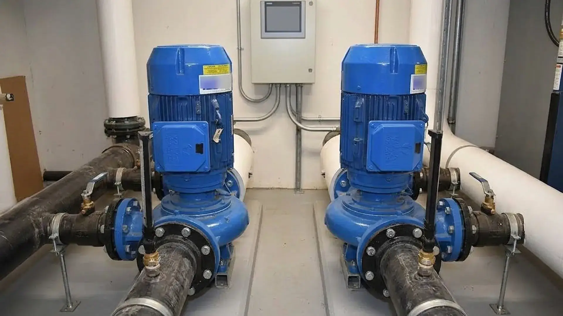 Two vertical inline pumps installed in parallel in an HVAC mechanical room with straight-through pipe routing, common headers, isolation valves, and a wall-mounted VFD panel, showing inline pump vs end suction pump space and maintenance trade-offs.